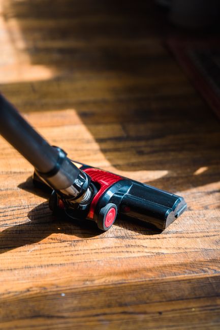 Close-up of a vacuum cleaner head lying on a wooden floor in a residential room, with sunlight casting shadows on the surface. The vacuum head is black and red, with a sleek design, and part of the metal extension tube is visible. The wooden floor shows a polished, clean appearance with distinct grain patterns and warm tones. This image illustrates surface cleaning and maintenance as part of domestic or deep cleaning services offered by Cleaners SW12, highlighting their attention to hygiene and detailed cleaning processes.
