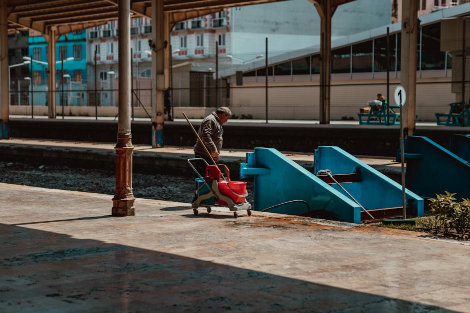 A man is standing on a concrete platform at Balham Station, holding a broom and facing towards the blue staircase leading down to the train tracks. The platform is partially shaded, with a lamp post in the foreground and a small bush visible on the right side. In the background, there are benches with a person sitting and the station’s modern glass and metal structures. The scene captures the urban environment of Balham Station, emphasizing the cleanliness and maintenance activities typical of a well-kept railway platform. This description aligns with the services provided by Cleaners SW12 for station and commercial cleaning, highlighting surface maintenance and hygiene standards.