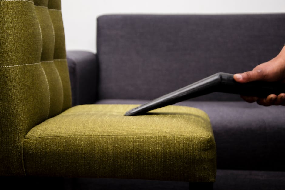 Close-up of a person using a handheld vacuum cleaner to clean a light green upholstered chair with a button-tufted backrest, in a room with a dark gray sofa in the background. The lighting highlights the clean surface of the fabric, indicating a thorough surface cleaning process. The scene is part of domestic cleaning services offered by Cleaners SW12, specializing in upholstery cleaning and stain removal in Bedford Hill, Balham, SW12. The focus is on the detailed cleaning of the fabric upholstery, showcasing the use of professional tools to maintain hygiene and appearance of furniture surfaces.