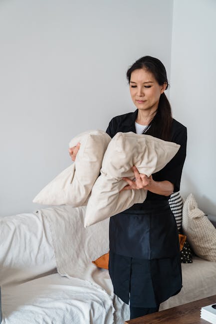 A professional cleaner from Cleaners SW12 is preparing to undertake upholstery cleaning or stain removal in a residential living room, holding two beige cushions. The room features a white sofa with a neutral-colored cushion and a beige textured throw on the armrest, positioned against a plain white wall. Natural light illuminates the space, highlighting the clean and tidy surface of the sofa, which appears free from dust or stains. The cleaner is dressed in a black uniform, and the setting suggests a focus on deep cleaning and maintaining hygiene within a domestic environment in line with Bedford Hill upholstery cleaning and stain removal services in Balham, SW12.