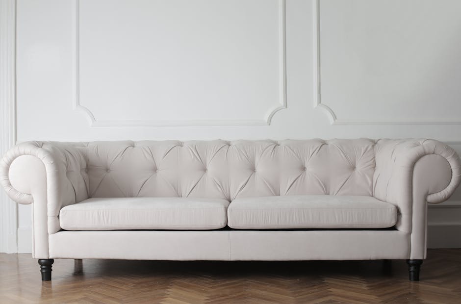 Close-up of a person using a handheld vacuum cleaner to clean a light green upholstered chair with a button-tufted backrest, in a room with a dark gray sofa in the background. The lighting highlights the clean surface of the fabric, indicating a thorough surface cleaning process. The scene is part of domestic cleaning services offered by Cleaners SW12, specializing in upholstery cleaning and stain removal in Bedford Hill, Balham, SW12. The focus is on the detailed cleaning of the fabric upholstery, showcasing the use of professional tools to maintain hygiene and appearance of furniture surfaces.
