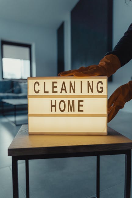 A person wearing orange gloves is placing their hand on a small illuminated sign on a wooden table, displaying the words 'CLEANING HOME' in black letters on a white background. The sign is in the fore
