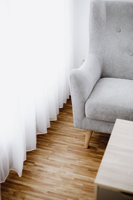 Close-up of a light grey upholstered armchair with wooden legs positioned in a well-lit room next to a sheer white curtain hanging beside a wooden floor. The fabric of the chair appears clean and smooth, with a visible tufted button detail on the backrest. Adjacent to the chair, a light-colored wooden side table is partially visible. The room has natural daylight filtering through the curtain, highlighting the pristine condition of the furniture and flooring. This setting exemplifies professional domestic surface cleaning aimed at maintaining hygiene and a tidy appearance, as offered by Cleaners SW12 for their upholstery cleaning services in Tooting Bec Common, SW12.