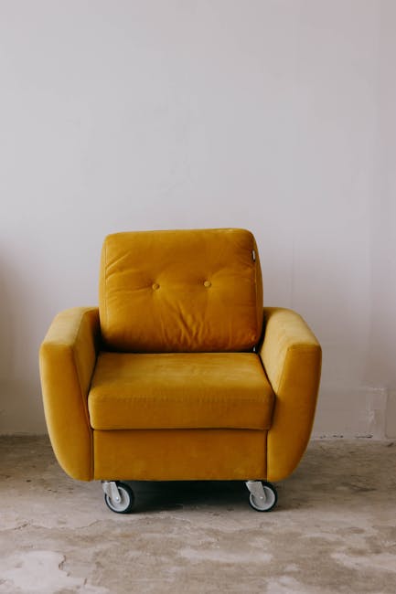 A mustard yellow armchair with a plush, tufted backrest and wide armrests, positioned on a concrete floor against a plain white wall. The chair is on small castor wheels, indicating it may be mobile or part of a cleaning or maintenance process. The surface of the upholstery appears clean and well-maintained, reflecting a tidy, freshly cleaned condition in a modern indoor setting. The lighting is natural, subtly illuminating the room without harsh shadows, consistent with a domestic or commercial space. This image exemplifies surface cleaning and care, as promoted by Cleaners SW12 in their upholstery cleaning services for properties around Tooting Bec Common, Balham, SW12.