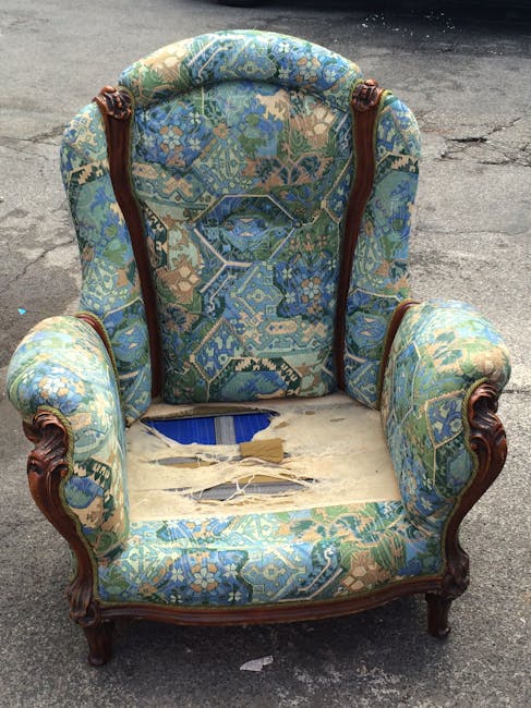Close-up of a light grey upholstered armchair with wooden legs positioned in a well-lit room next to a sheer white curtain hanging beside a wooden floor. The fabric of the chair appears clean and smooth, with a visible tufted button detail on the backrest. Adjacent to the chair, a light-colored wooden side table is partially visible. The room has natural daylight filtering through the curtain, highlighting the pristine condition of the furniture and flooring. This setting exemplifies professional domestic surface cleaning aimed at maintaining hygiene and a tidy appearance, as offered by Cleaners SW12 for their upholstery cleaning services in Tooting Bec Common, SW12.